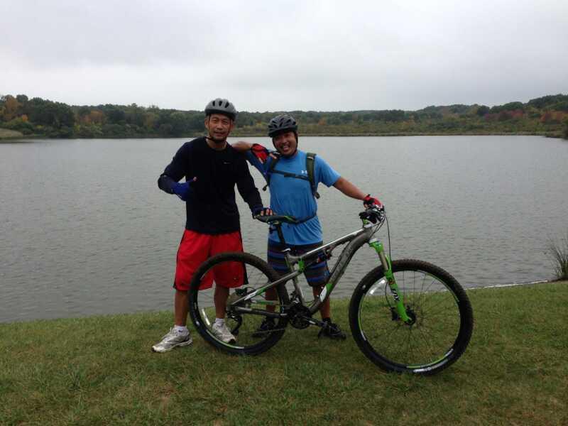 Two men pose together with a mountain bike beside a calm lake. One man is wearing a black shirt and red shorts, while the other is in a blue shirt and colorful shorts. They both wear helmets and smile at the camera, with green trees in the background under a cloudy sky. Palos Forest Preserve mountain bike trail.