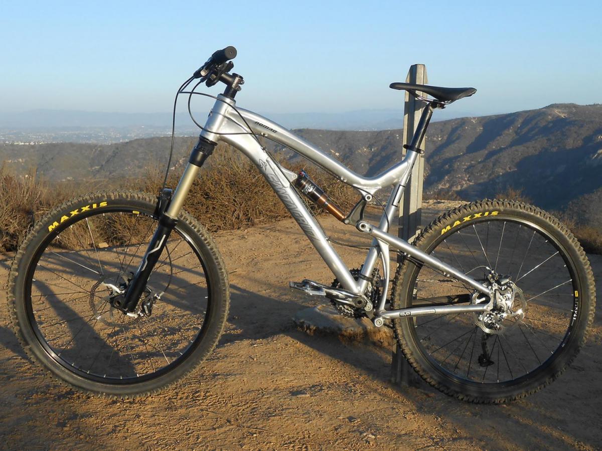 Santa Cruz Nomad: A silver mountain bike is parked on a dirt trail with scenic views of distant mountains and valleys in the background. The bike features thick tires and a sturdy frame, positioned next to a wooden post. The image captures a clear sky, suggesting a bright and sunny day for outdoor biking.