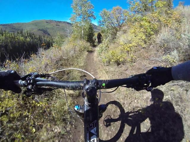A perspective view from a mountain bike handlebars, showcasing a dirt trail winding through a vibrant outdoor landscape filled with trees and foliage. In the background, a cyclist can be seen riding along the trail under a clear blue sky. Park City Mountain Resort (PCMR) mountain bike trail.