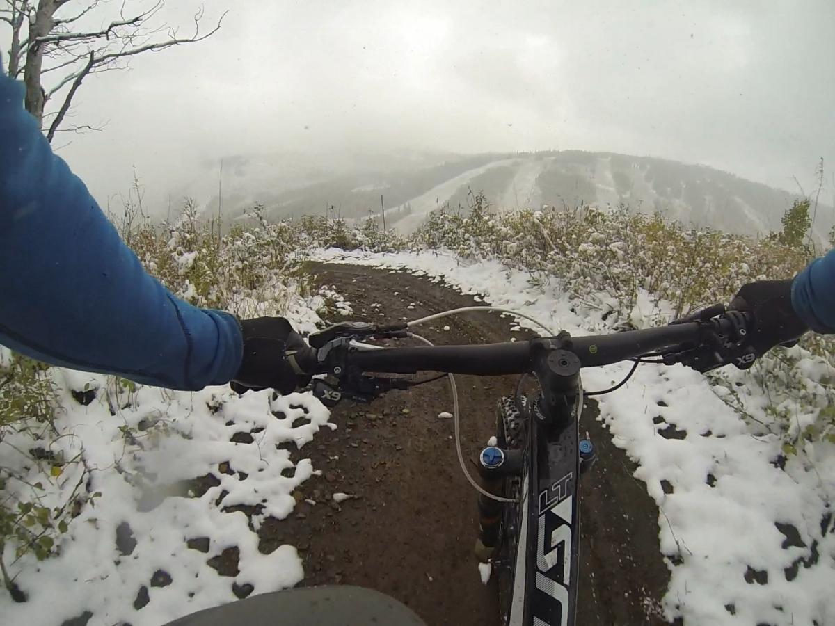 Mountain biker's perspective on a snowy trail, with hands on the handlebars and a view of a winter landscape featuring snow-covered ground and distant hills. Pinecone Ridge mountain bike trail.