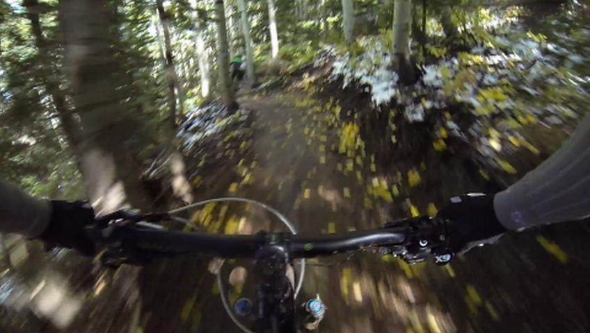 A close-up view of a mountain bike handlebar navigating a dirt trail surrounded by trees, with a foreground showing blurred yellow autumn leaves scattered on the ground. The background features a cyclist in green clothing riding along the trail. The image conveys a sense of speed and adventure in a natural setting. Park City Mountain Resort (PCMR) mountain bike trail.