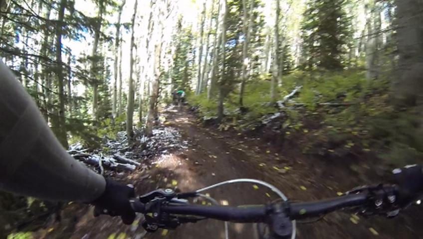 A mountain biking perspective shot on a dirt trail surrounded by tall trees, with patches of sunlight filtering through the leaves. The handlebars of the bicycle are visible in the foreground, and a blurred figure riding a bike can be seen further down the trail. Some areas of the trail have fallen leaves and light snow. Park City Mountain Resort (PCMR) mountain bike trail.