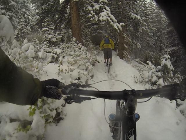 Two mountain bikers ride along a snow-covered trail through a forest. The foreground shows the handlebars of one bike, while the other biker, wearing a yellow jacket, is visible in the background navigating the snowy path surrounded by tall trees. Pinecone Ridge mountain bike trail.