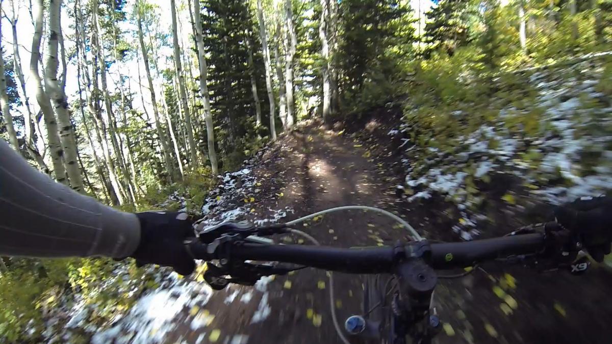 A view from the handlebars of a mountain bike riding along a narrow dirt trail surrounded by trees. The ground shows patches of snow and fallen leaves, indicating a transition between seasons. Sunlight filters through the trees, creating dappled light effects on the trail. Park City Mountain Resort (PCMR) mountain bike trail.