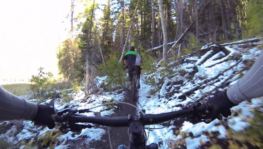A mountain biking scene showcasing a person's perspective from the handlebars, riding along a narrow, snowy trail surrounded by trees. Another biker is visible ahead, navigating the path through the forest. The sunlight filters through the trees, highlighting the natural landscape. Park City Mountain Resort (PCMR) mountain bike trail.