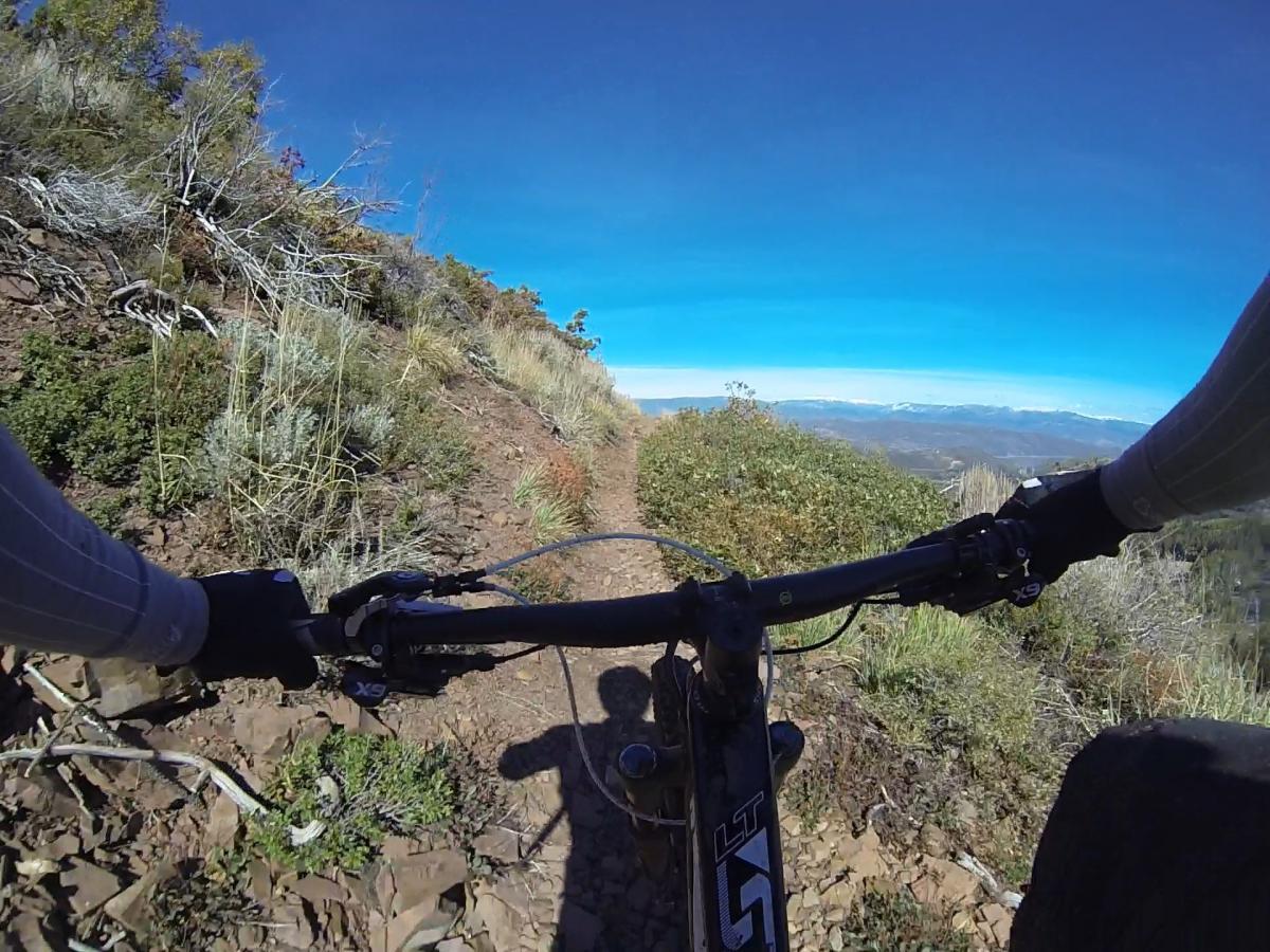A mountain biking perspective showing the handlebars of a bike on a narrow trail surrounded by green vegetation, with a blue sky and distant mountains visible in the background. Park City Mountain Resort (PCMR) mountain bike trail.