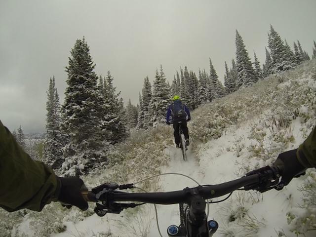 A mountain biking scene featuring two cyclists navigating a snowy trail surrounded by evergreen trees under a cloudy sky. The view is taken from the handlebars of the bike in the foreground, showcasing the snowy path ahead and the cyclist in a blue jacket riding in front. Scott's Bypass mountain bike trail.