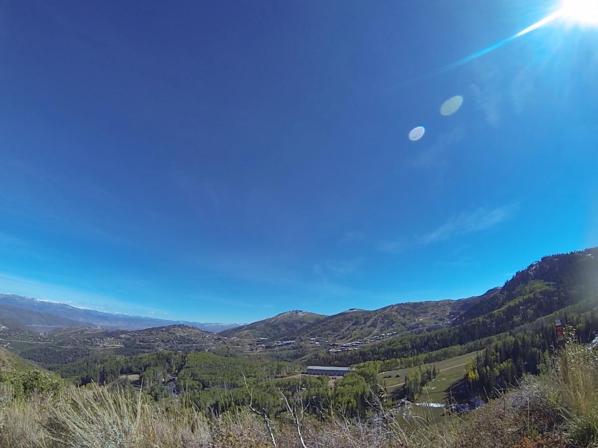 A panoramic view of a mountainous landscape under a clear blue sky, featuring rolling hills covered with lush green trees, a small valley, and distant snowy peaks. The sunlight creates a bright glare in the top right corner of the image. Park City Mountain Resort (PCMR) mountain bike trail.