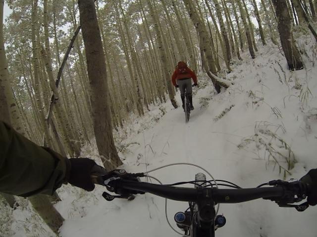 A first-person perspective of mountain biking through a snowy forest, with a cyclist in a red jacket riding ahead on a narrow path lined with trees. Snow covers the ground, creating a winter landscape. Scott's Bypass mountain bike trail.