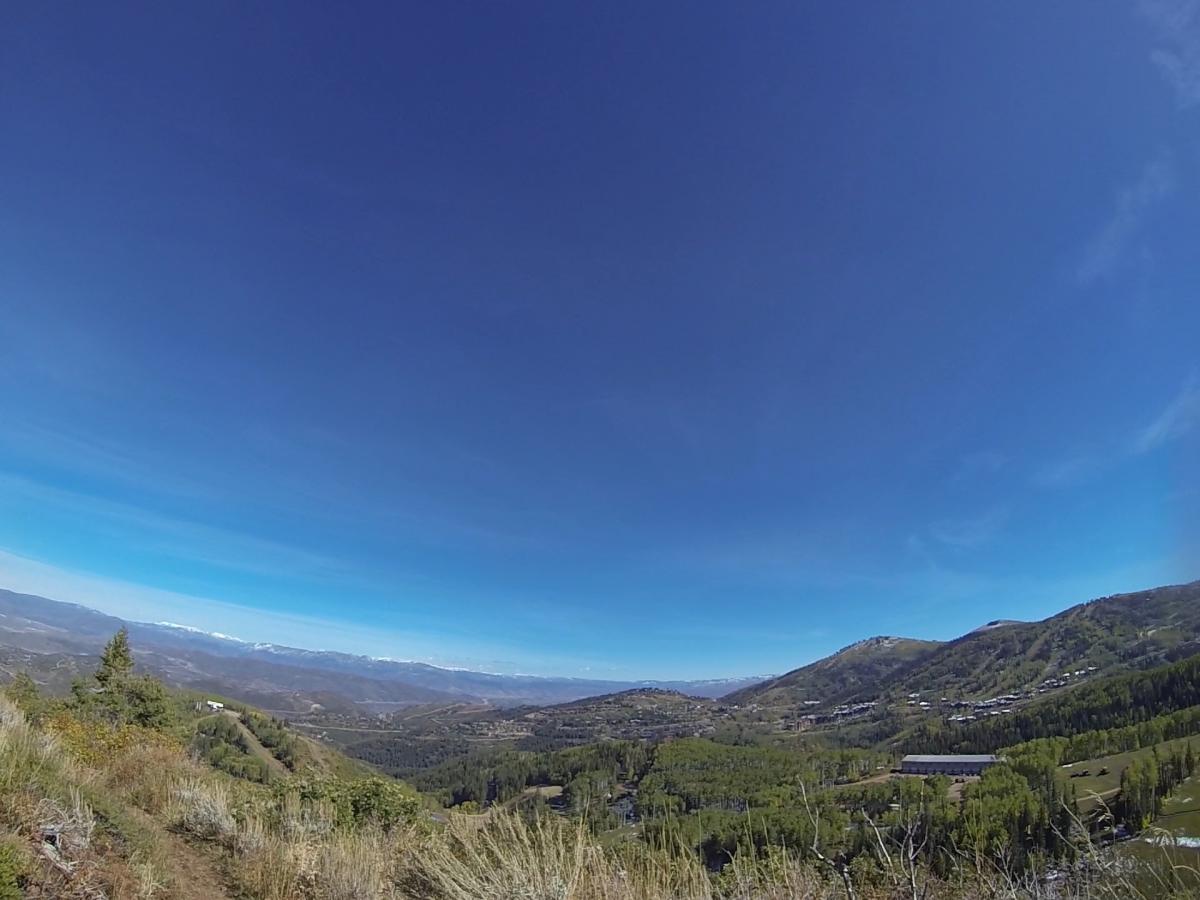 A panoramic view of a mountainous landscape under a clear blue sky, with lush green hills and distant snow-capped peaks. The foreground features grass and shrubs, while valleys and small structures are visible in the mid-ground. Park City Mountain Resort (PCMR) mountain bike trail.