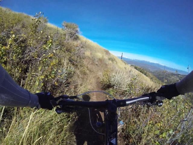 A first-person view of a mountain bike handlebar navigating a narrow dirt trail surrounded by tall grass and small shrubs, with a clear blue sky and distant mountains visible in the background. Park City Mountain Resort (PCMR) mountain bike trail.
