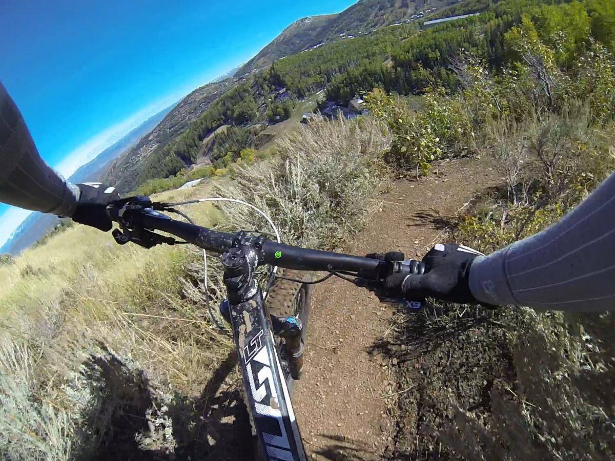 A view from a mountain bike descending a rugged trail, with the handlebars in focus and hands wearing gloves. The landscape features lush greenery and rolling hills under a clear blue sky. Park City Mountain Resort (PCMR) mountain bike trail.