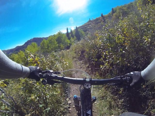 A view from the handlebars of a mountain bike, showcasing a narrow dirt trail surrounded by lush greenery and trees, with a clear blue sky above. The image conveys the feeling of riding through a scenic outdoor landscape. Park City Mountain Resort (PCMR) mountain bike trail.