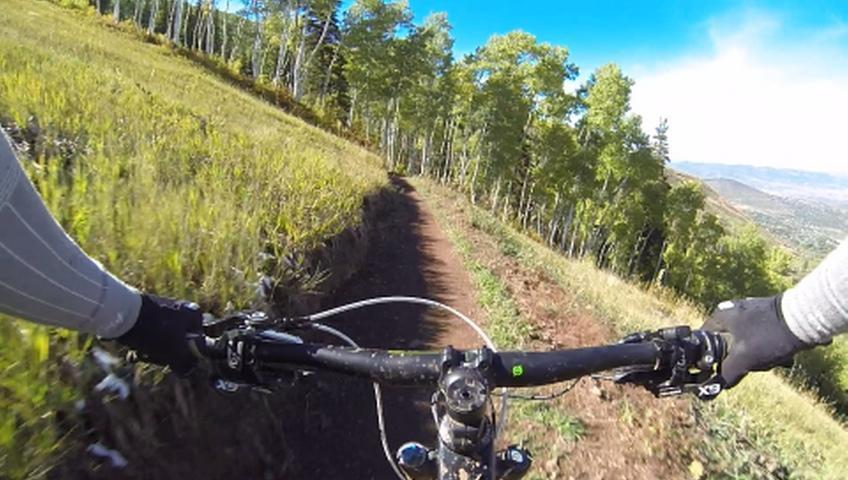 Alt text: A first-person view of a mountain biking trail, showing handlebars and a winding dirt path surrounded by lush green grass and trees under a clear blue sky. Park City Mountain Resort (PCMR) mountain bike trail.