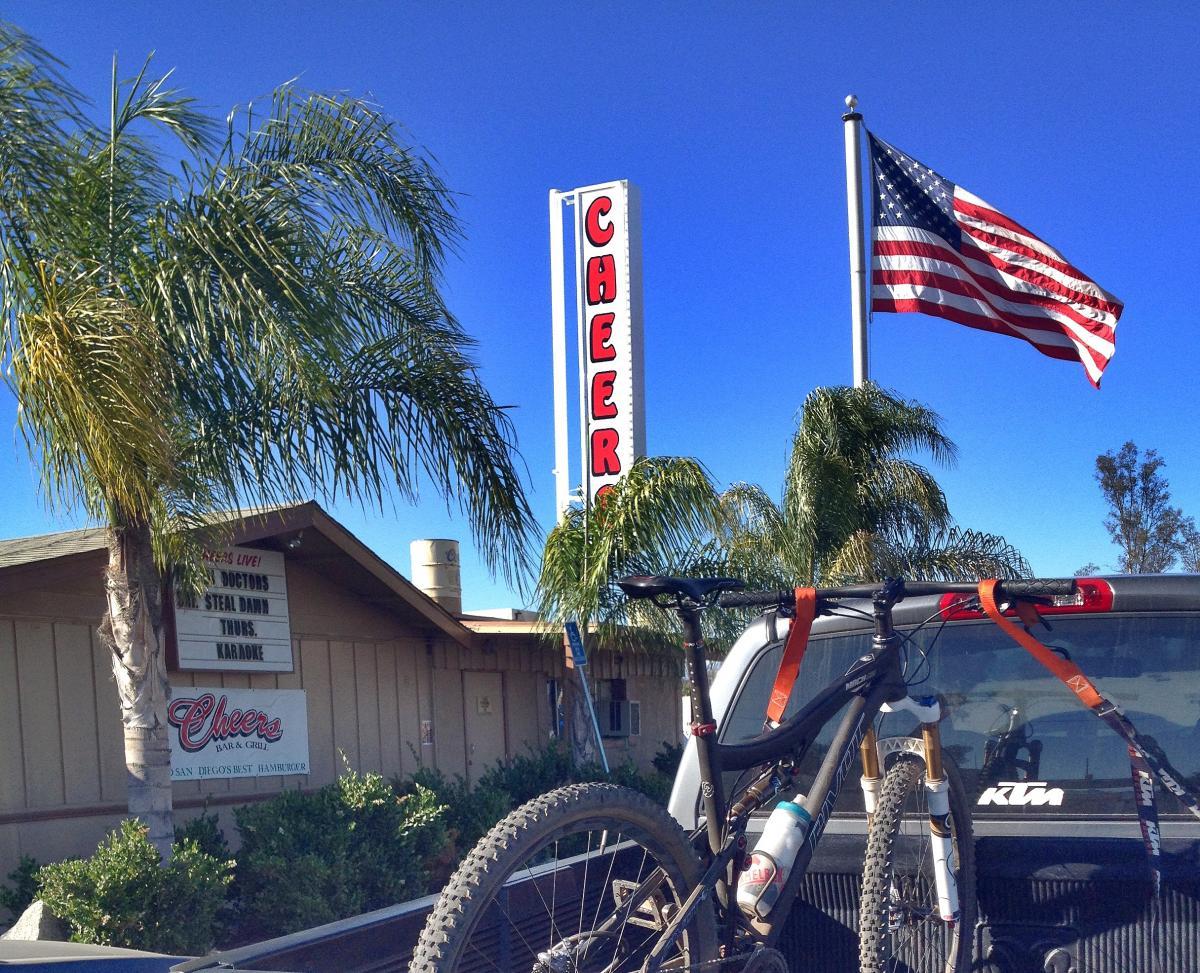 Pivot Mach 429: A blue sky above a roadside bar and grill named "Cheers," featuring a bright red sign. In the foreground, a mountain bike is secured in the back of a truck, with orange straps holding it in place. Nearby, a flagpole displays the American flag, and palm trees frame the scene. A sign on the building shows upcoming events like karaoke.