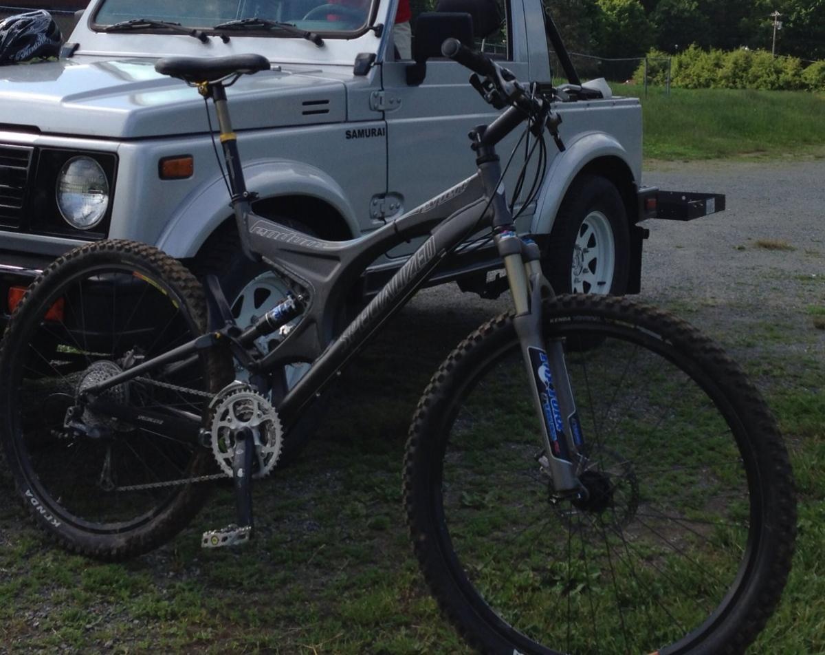 Specialized Enduro Expert: A mountain bike with a sturdy frame and thick tires is positioned in front of a silver Suzuki Samurai. The bike features a suspension system and a chain system visible near the rear wheel. The background consists of grass and a partially visible blue sky.