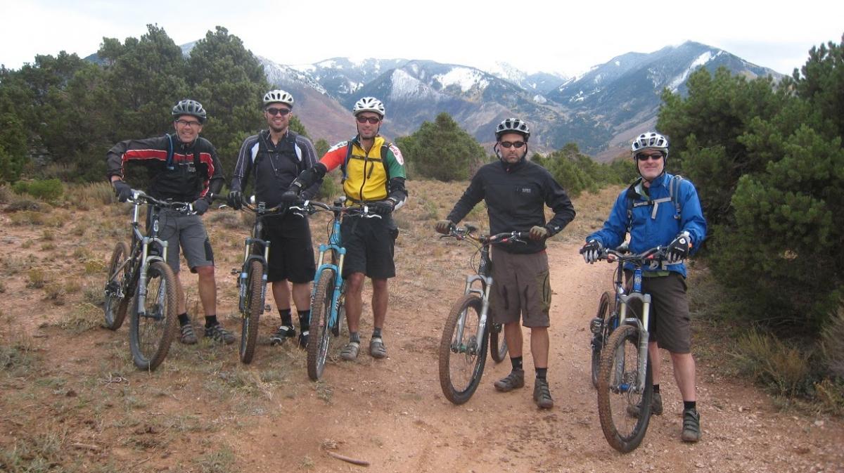 Five mountain bikers pose together on a dirt trail surrounded by greenery and mountains in the background. They are wearing cycling gear and helmets, with their bikes beside them. The scene suggests an outdoor adventure in a mountainous area. Porcupine Rim mountain bike trail.