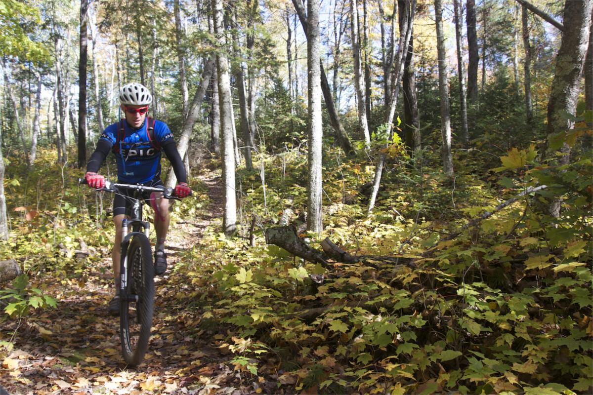 A person riding a mountain bike along a wooded trail, surrounded by autumn foliage. The cyclist is wearing a blue jersey with "EPIC" printed on it, along with a helmet and sunglasses. Sunlight filters through the trees, highlighting the colorful leaves on the ground. Rock Lake mountain bike trail.