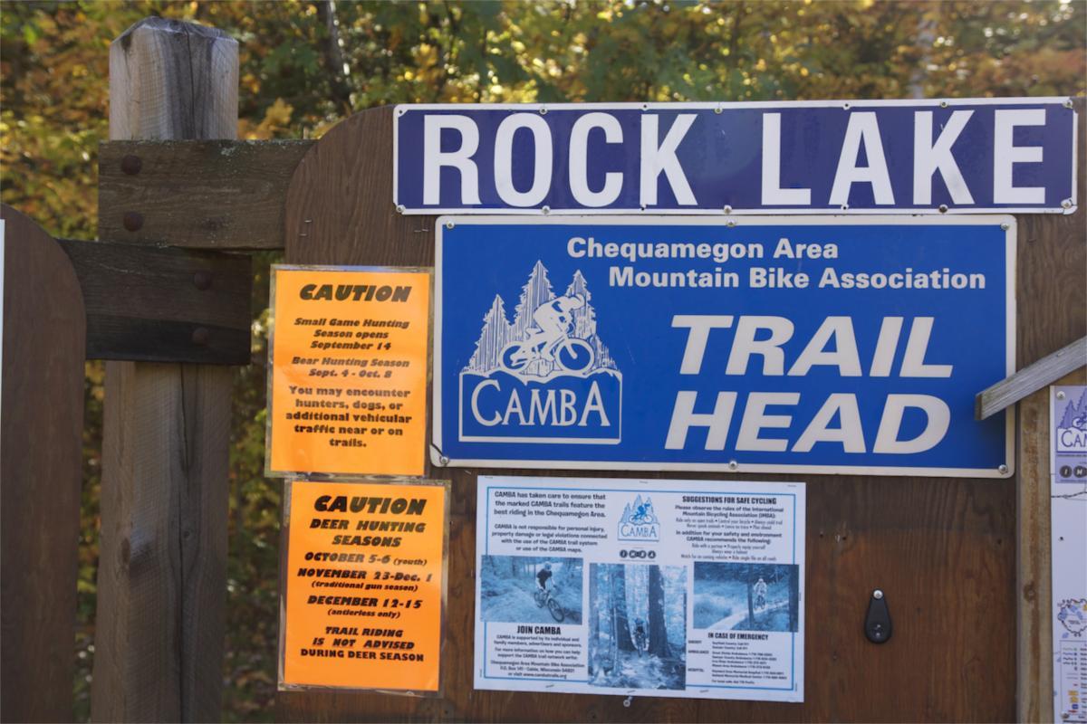 Signage at the Rock Lake trailhead, displaying information about the Chequamegon Area Mountain Bike Association (CAMBA). The signs include cautions for small game and bear hunting seasons, as well as deer hunting seasons, with specific dates. Additional notes mention the potential for encountering hunters and vehicles on the trails, along with safety suggestions for cyclists. Rock Lake mountain bike trail.