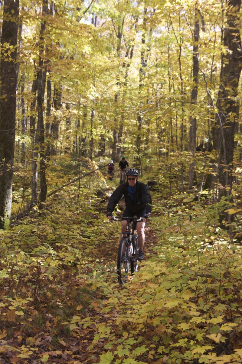 A person riding a mountain bike on a narrow trail surrounded by vibrant autumn foliage in a dense forest. The ground is covered with fallen leaves, and more cyclists can be seen in the background. Rock Lake mountain bike trail.