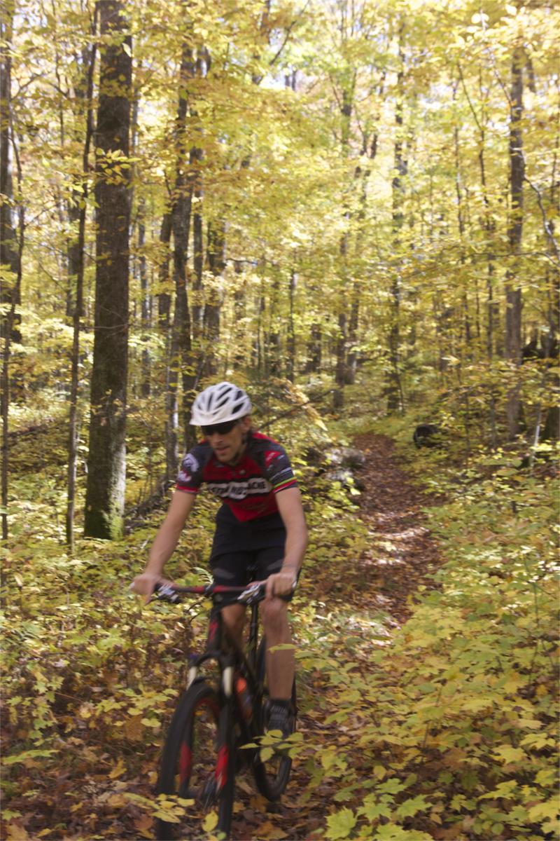 A cyclist riding a mountain bike on a dirt path through a vibrant autumn forest filled with yellow and orange leaves. Rock Lake mountain bike trail.