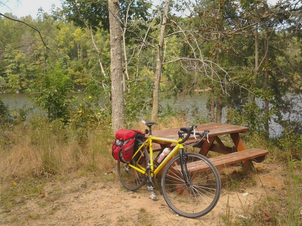 A bright yellow bicycle with a red saddlebag is parked beside a wooden picnic table near a river, surrounded by greenery and trees. Salem Lake mountain bike trail.