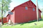 A red barn with a sloped roof, partially obscured by trees. The building features windows on the upper level and a sign hanging near the entrance. Green grass surrounds the barn, and a utility pole is visible in the background.