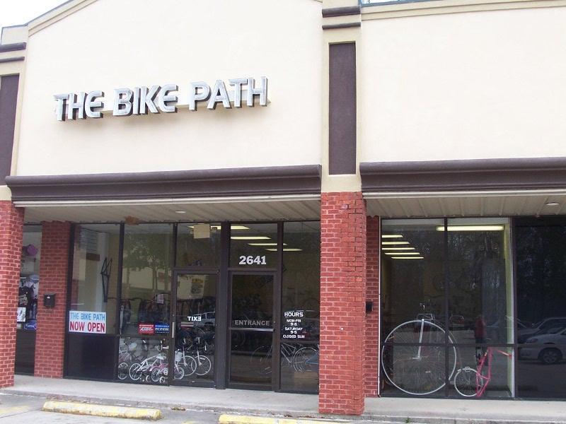 Front view of a bike shop named "The Bike Path," featuring large windows displaying bicycles inside. The sign above the entrance indicates "NOW OPEN," and there is a visible address (2641) next to the door. The storefront has a mix of light-colored and red brick elements.