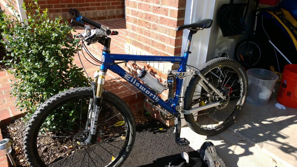 A blue mountain bike with a silver frame is parked on a driveway next to a bush. The bike features visible suspension components, a water bottle holder, and knobby tires. In the background, there's a brick wall and various items stored in a garage, including a shovel and a bike trailer.