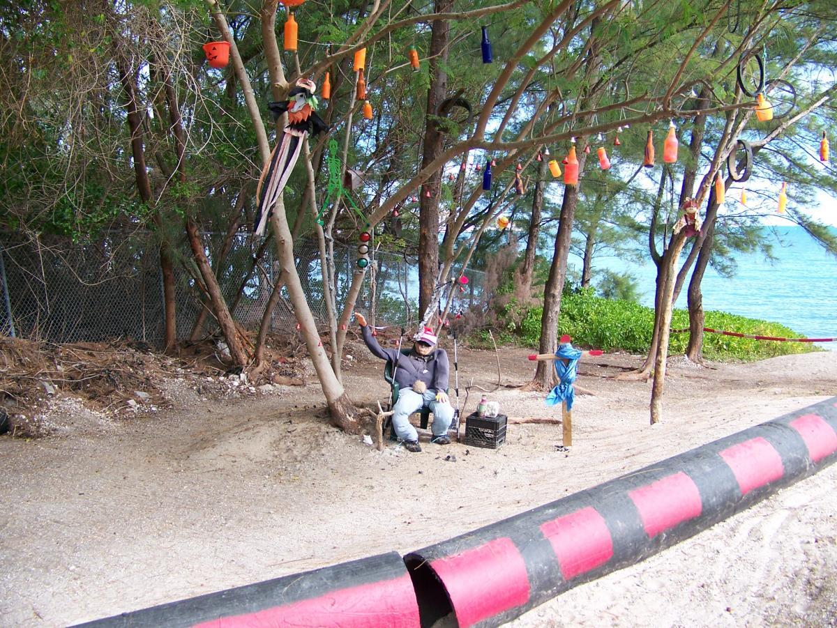 A sandy pathway lined with trees, featuring colorful decorations hanging from the branches. Two figures are seated, one dressed casually with a hat, appearing to relax amidst the scenic backdrop of the ocean. The foreground includes a black and red striped barrier. The overall setting conveys a tranquil beach atmosphere. Virginia Key North Point mountain bike trail.