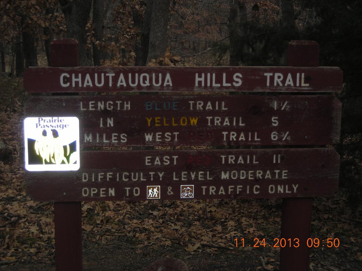 Sign for the Chautauqua Hills Trail, displaying trail lengths for the blue, yellow, and red trails. The sign indicates the blue trail is 1.5 miles, the yellow trail is 5 miles, and the red trail is 6.75 miles. It also notes the difficulty level is moderate and that the trail is open to foot traffic and bicycles. In the background, trees and fallen leaves can be seen. Chautauqua Hills Trail mountain bike trail.