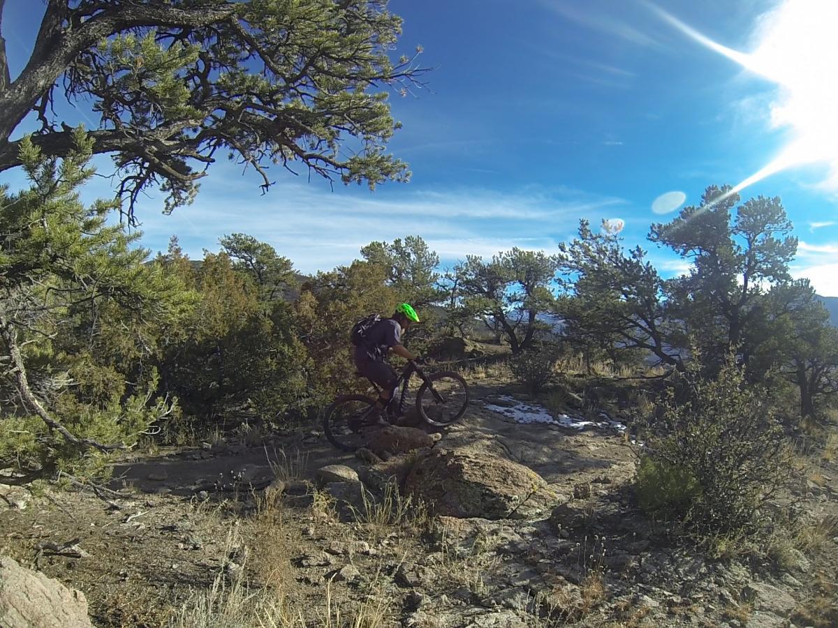 A mountain biker navigates over rocky terrain surrounded by trees and a clear blue sky. The cyclist is wearing a bright green helmet and dark clothing, demonstrating an active outdoor adventure in a natural setting. Unkle Nazty mountain bike trail.