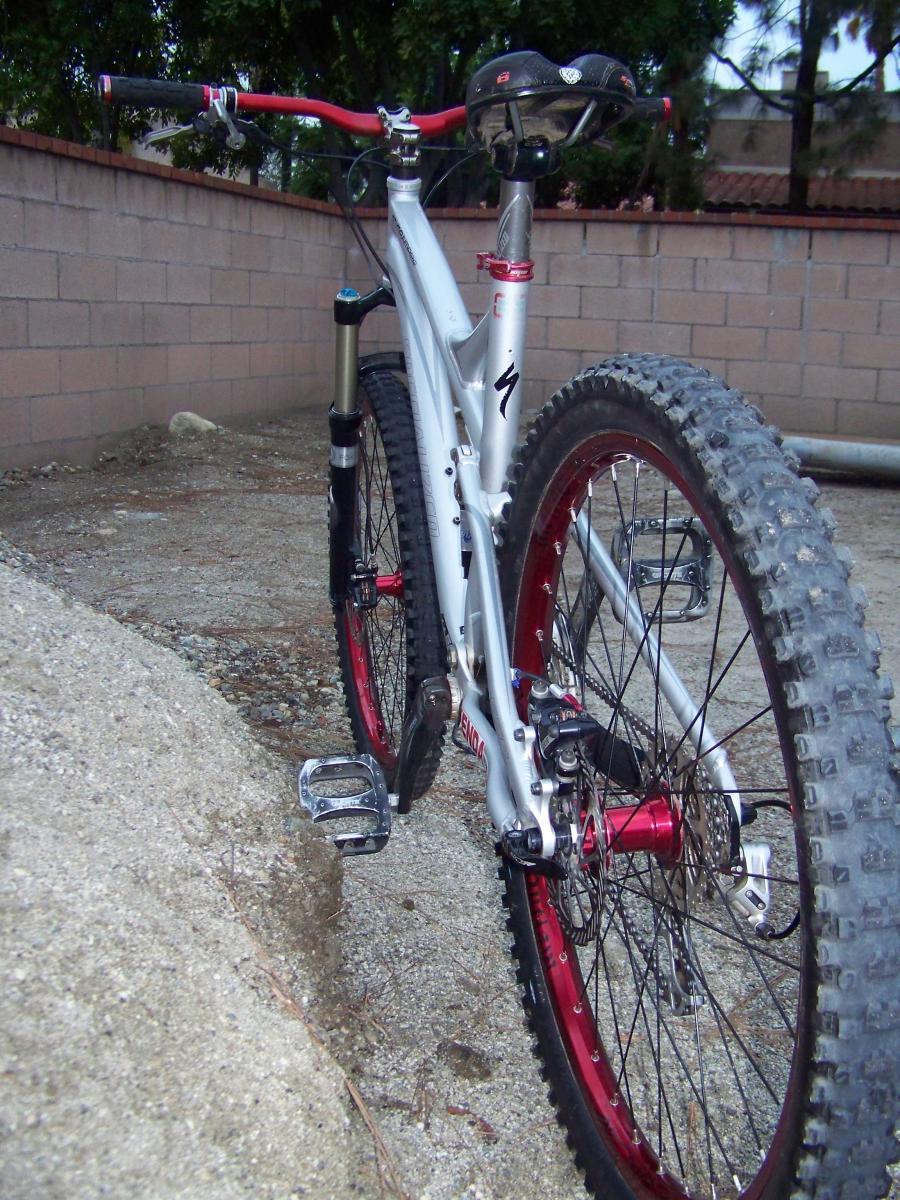 Specialized Stumpjumper: Alt text: A close-up view of a silver mountain bike with red accents, showcasing the rear tire, pedals, and part of the frame. The background features a gravel surface and a brick wall.