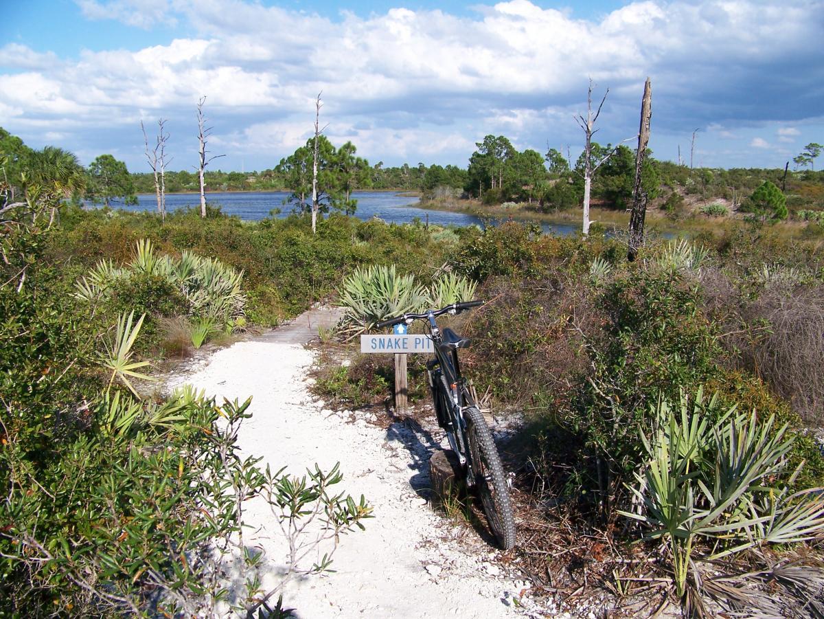 A mountain bike leaning against a sign marked "Snake Pit" on a sandy pathway surrounded by lush greenery, leading to a serene lake with trees in the background under a partly cloudy sky. Jonathan Dickinson State Park mountain bike trail.