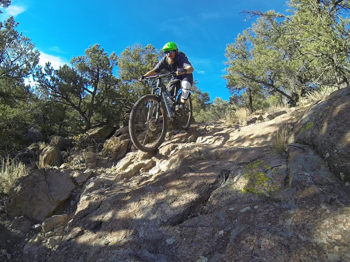 A mountain biker navigating rocky terrain in a forested area, wearing a helmet and protective gear, with trees and blue skies visible in the background. Unkle Nazty mountain bike trail.
