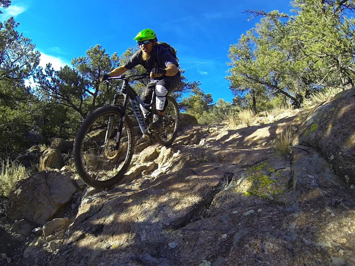 A mountain biker with a green helmet navigates a rocky trail surrounded by trees and blue sky. The cyclist is mid-action, riding over uneven terrain, showcasing a dynamic outdoor adventure in nature. Unkle Nazty mountain bike trail.