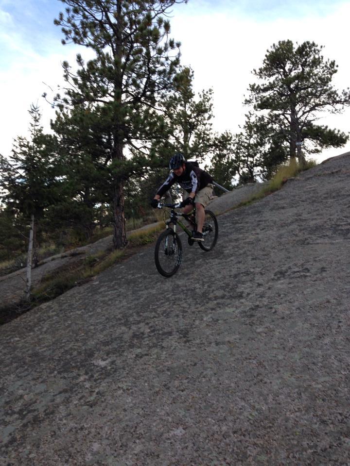 Cannondale RZ 120-1: A mountain biker riding downhill on a rocky trail surrounded by trees, with a clear blue sky in the background. The cyclist is dressed in a helmet and protective gear, demonstrating an adventurous descent on the uneven terrain.