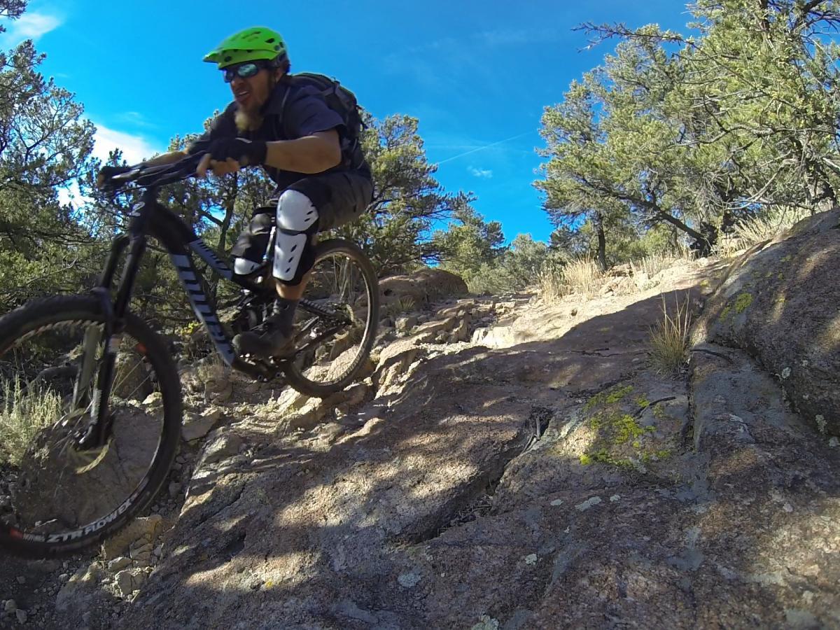 A mountain biker wearing a green helmet and protective gear navigates a rocky trail surrounded by trees under a bright blue sky. The cyclist is mid-jump, showcasing an adventurous spirit in an outdoor setting. Unkle Nazty mountain bike trail.
