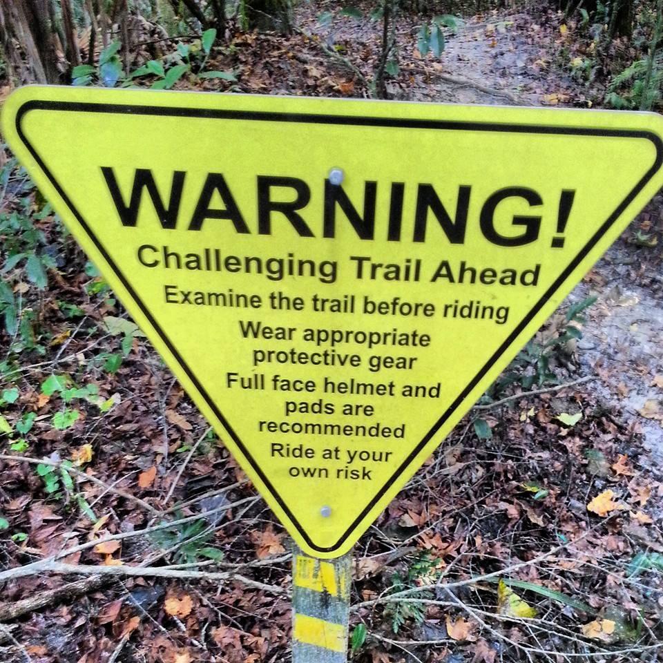 A yellow triangular warning sign indicating a challenging trail ahead, advising riders to examine the trail before riding, wear appropriate protective gear, and recommending the use of a full-face helmet and pads. The sign warns to ride at your own risk. Surrounding the sign are trees and fallen leaves, suggesting a forested area. Chuck Lennon Park mountain bike trail.