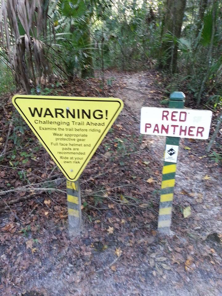 Image of two signs at the entrance of a trail. The left sign, yellow and triangular, warns of a "Challenging Trail Ahead," advising riders to examine the trail, wear appropriate protective gear, and emphasizes that riding is at one's own risk. The right sign indicates the trail name, "RED PANTHER." The surrounding area features dense foliage and a dirt path. Chuck Lennon Park mountain bike trail.