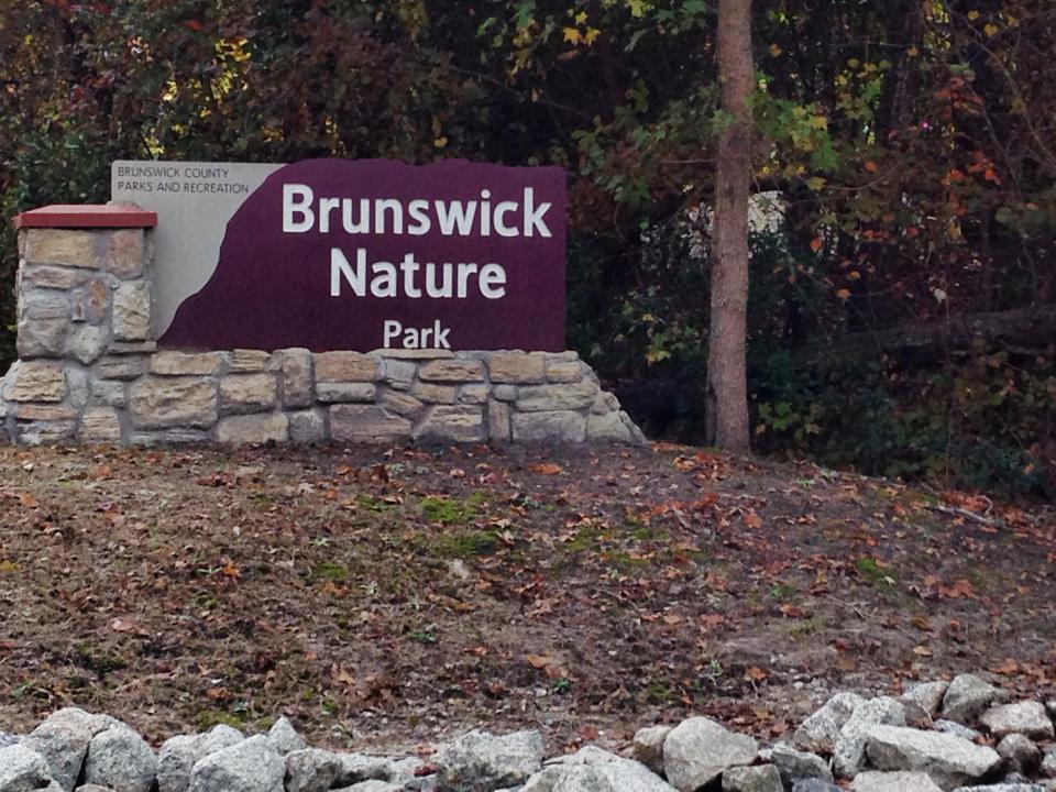 Sign for Brunswick Nature Park, featuring stone and wood elements, surrounded by trees and foliage. The sign includes text that states "Brunswick County Parks and Recreation" and "Brunswick Nature Park" prominently. Brunswick Nature Park mountain bike trail.