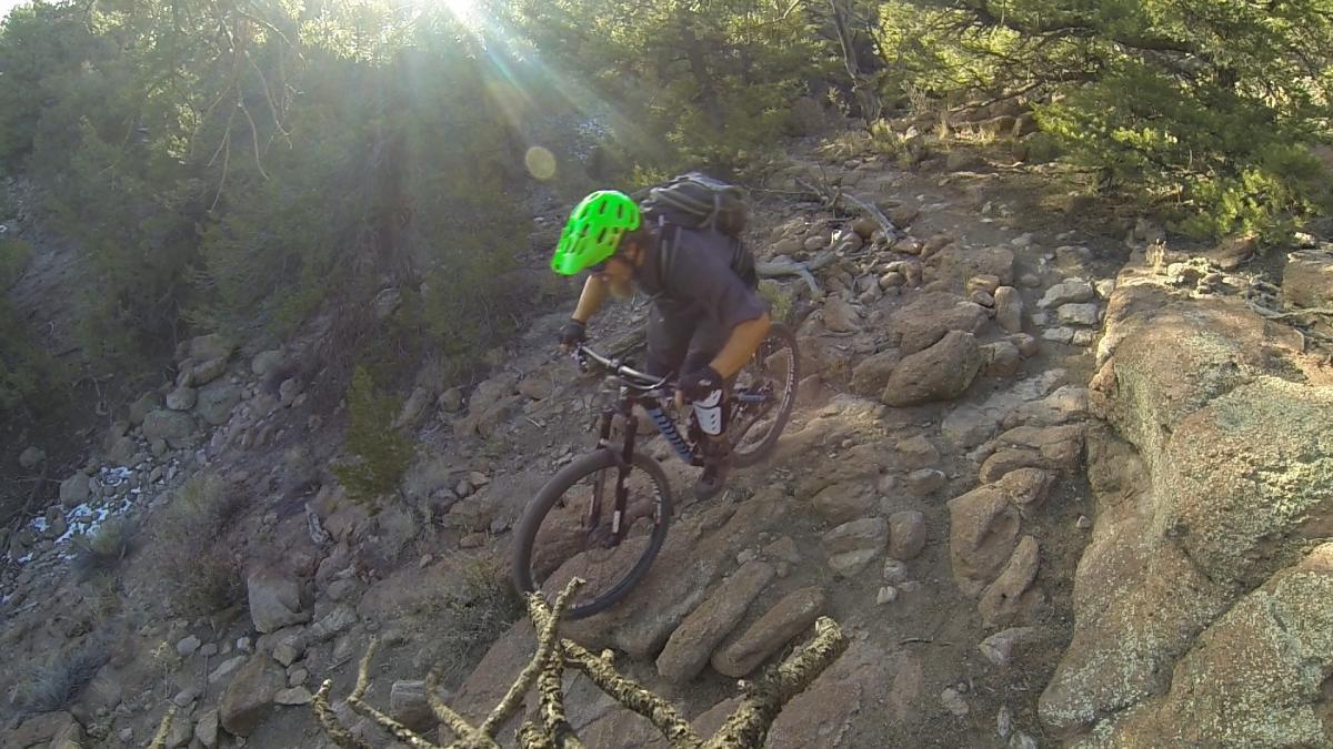 A mountain biker navigates a rocky trail surrounded by trees, wearing a bright green helmet and a backpack. The sun shines through the foliage, highlighting the rugged terrain. Unkle Nazty mountain bike trail.