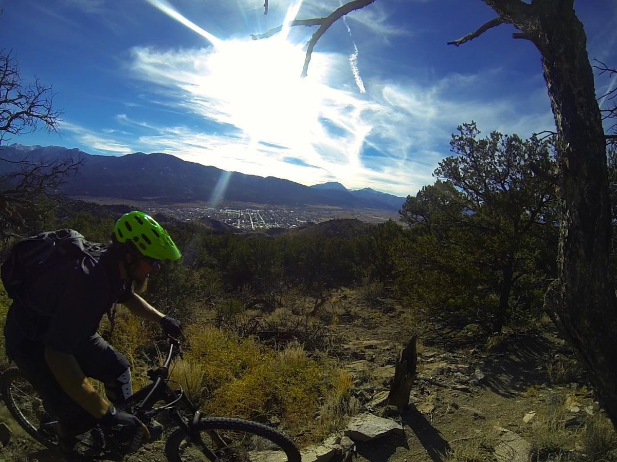 A mountain biker wearing a bright green helmet navigates a rocky trail with a mountainous landscape in the background. The sun shines brightly in a blue sky with wispy clouds, illuminating the scene. Below, a small town is visible nestled in the valley. Unkle Nazty mountain bike trail.