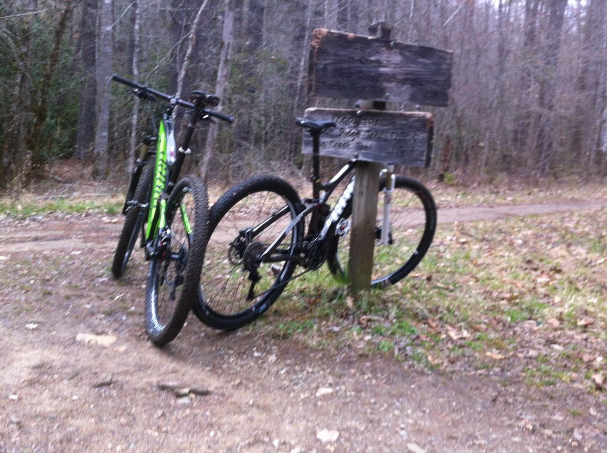 Two mountain bikes are leaning against a wooden trail sign in a wooded area. The sign has faded text and arrows indicating directions. The ground is a mix of dirt and scattered leaves, surrounded by trees in the background.