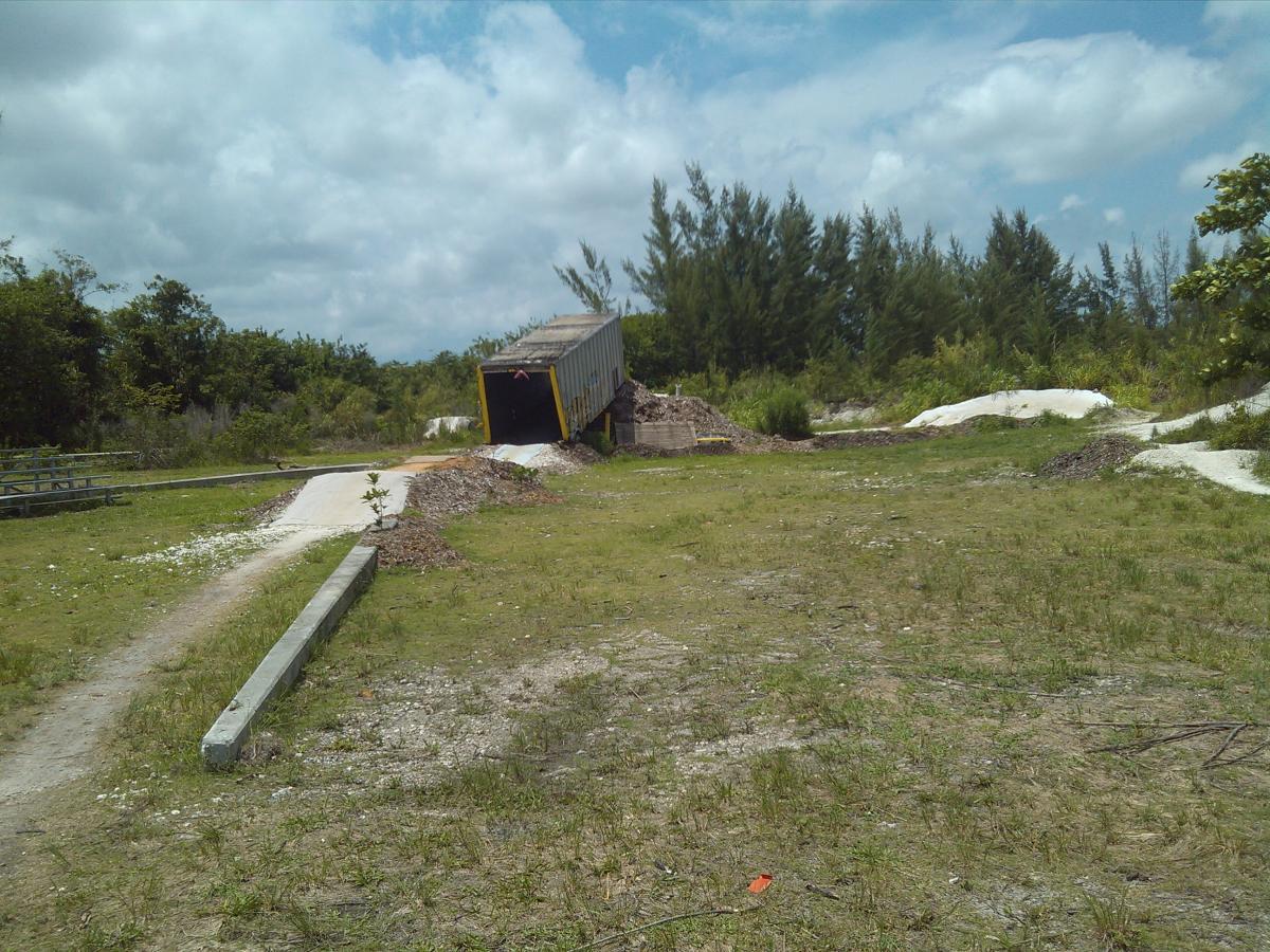 A shipping container partially overturned on a grassy area, surrounded by trees and scattered debris. The sky is partly cloudy, and a dirt path leads towards the container, while a small plant grows nearby. Amelia Earhart Park mountain bike trail.