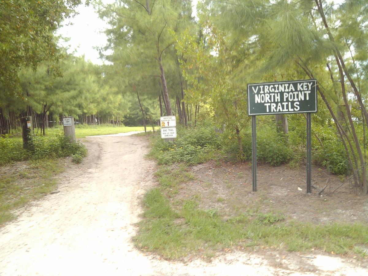 A dirt path leading into a wooded area with tall trees on either side. A green sign reads "Virginia Key North Point Trails," indicating the beginning of the trails. Additional signs are visible, providing information about trail rules and usage. Virginia Key North Point mountain bike trail.