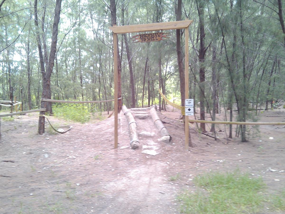 A wooden sign reading "Highway to Hell" marks the entrance to a trail in a forested area. The path is surrounded by tall trees and has logs resting on the ground. A warning sign indicates that the trail is for advanced users. The terrain is sandy with patches of grass and greenery nearby. Virginia Key North Point mountain bike trail.