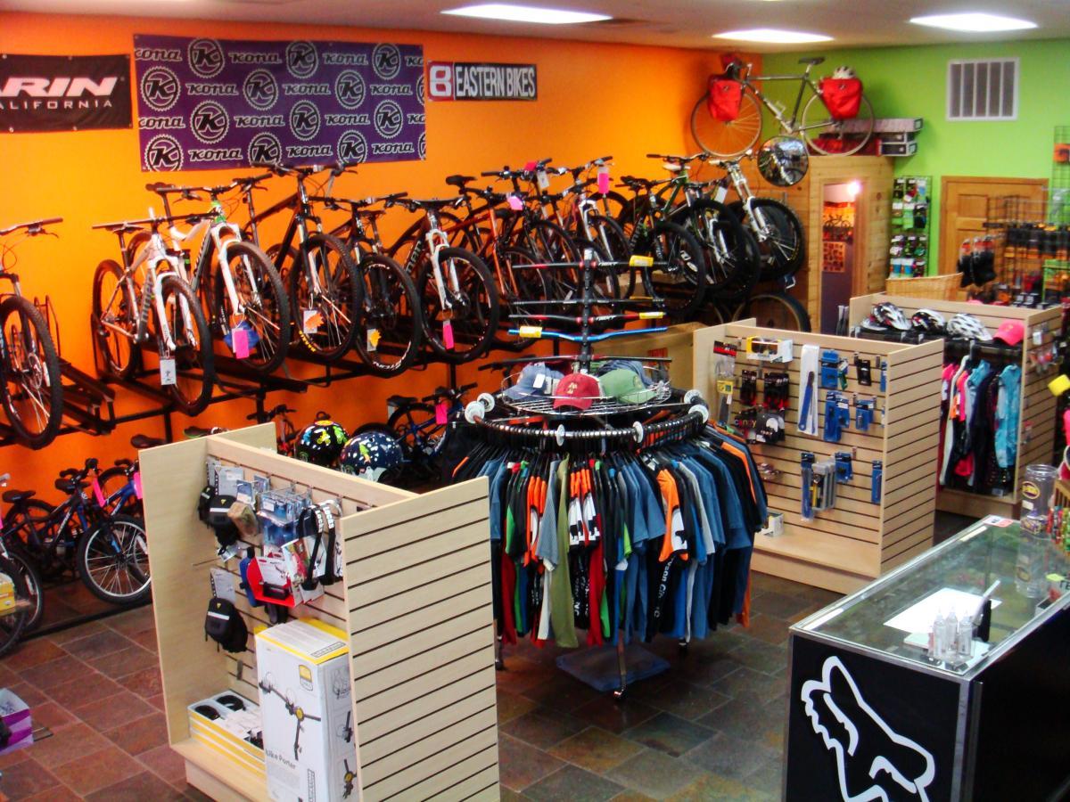 An indoor bike shop displaying a variety of bicycles mounted on orange walls. The store features a circular rack of colorful cycling apparel and accessories, alongside shelves filled with helmets and cycling gear. In the background, additional bikes are showcased, highlighting a vibrant and inviting shopping environment for cycling enthusiasts.