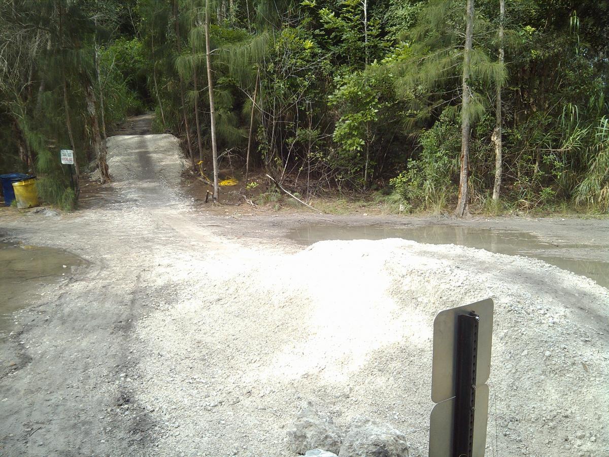 A dirt path diverges into two directions, surrounded by dense greenery. A pile of gravel is visible in the foreground, with a small body of water nearby. A sign post stands to the right, partially obscured, alongside a blue trash bin. The scene conveys a natural, unpaved road leading into a wooded area. Markham Park mountain bike trail.