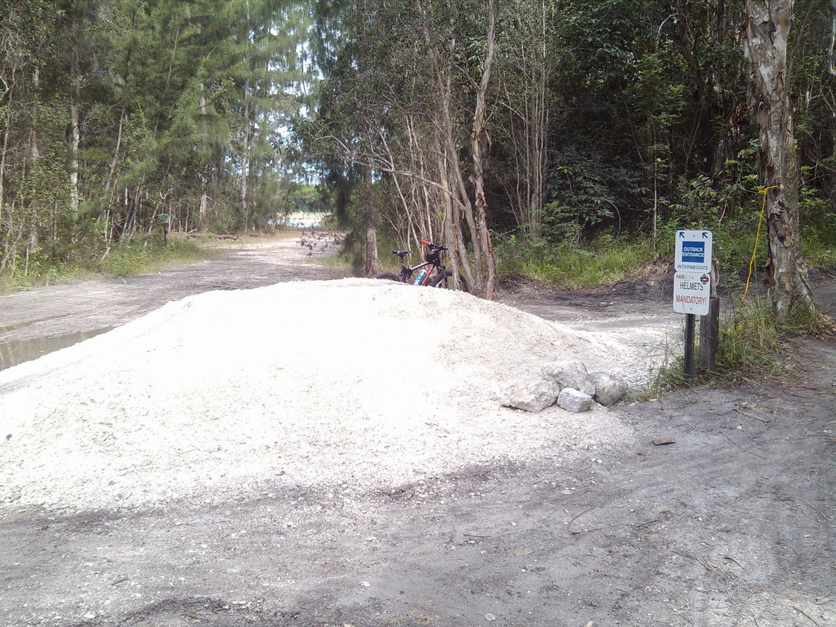 A dirt trail in a forested area, featuring a large pile of gravel at the foreground. A bicycle is parked near the gravel pile, and a sign on the right indicates trail directions and states that wearing helmets is mandatory. Trees line both sides of the path, creating a natural setting. Markham Park mountain bike trail.